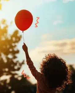 Niña de pelo rizado de espaldas a nosotros, en un parque. Tiene un globo rojo en la mano y su brazo se encuentra totalmente estirado. Es justo el momento antes de soltarlo, es decir una metáfora de despegarse emocionalmente de ese globo.