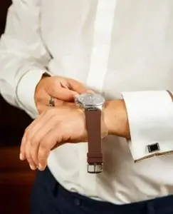 Hombre elegante, vestido con camisa blanca, gemelos y pantalones de traje azul oscuro. Justo en el momento de la foto se está colocando el reloj, pero parece que se ha parado a pensar si vale la pena esperar a alguien que no está preparado para una relación.