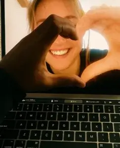 En esta imagen podemos ver la pantalla de ordenador portátil, en ella hay una chica en una sala iluminada por la luz del sol y haciendo un gesto con la mano. Enfrente de la pantalla se puede ver otra mano, del propietario del ordenador portátil, haciendo un gesto con la mano. Podemos ver que la unión de las dos manos genera el gesto del corazón. Parece que estos dos personajes se encuentran en una videollamada, y por el gesto, nos damos cuenta que a pesar de encontrarse a una gran distancia están en un buen momento de las etapas de una relación a distancia.
