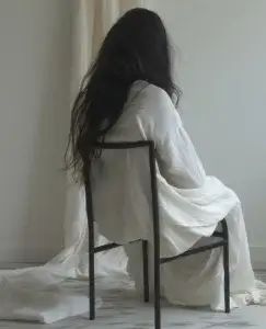 Mujer de pelo negro, sentada en una silla de hierro negro de espaldas a nosotros, esta vestida de blanco con un vestido largo y liso. Se encuentra en una habitación blanca, en la que hay una cortina blanca que nos recuerda su vestido. Está triste, se le nota por su postura caída y esto es porque está pensando en que su pareja no le apoya en sus momentos difíciles, por eso está sola y triste.