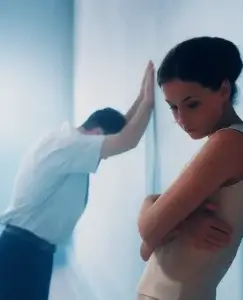 Una pareja aparece en una habitación iluminada en tonos fríos: ella está de espaldas, abrazándose a sí misma con expresión dolida, mientras él se inclina hacia la pared con gesto de frustración. La escena refleja la tensión y la dualidad emocional propias de una relación marcada por el amor-odio.