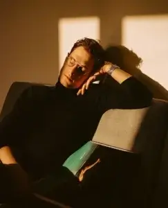 Un hombre joven descansa en un sofá iluminado por la luz cálida del atardecer, con los ojos cerrados y un libro abierto sobre sus piernas que muestra una fotografía en tonos verdes. La escena transmite una mezcla de calma y vulnerabilidad, como si estuviera procesando ese desconcierto silencioso que aparece cuando tu pareja te deja de golpe, quedándose por un momento suspendido entre el recuerdo y el intento de recuperar la serenidad.
