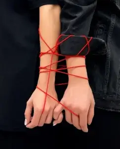 Dos brazos aparecen juntos, atados por varias vueltas de un hilo rojo que une firmemente sus muñecas, simbolizando una conexión que parece más restrictiva que afectiva. La imagen evoca la sensación de estar atrapado por una persona posesiva.