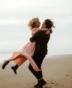 Una pareja se abraza en la playa mientras él levanta a ella con un gesto espontáneo de alegría; ambos sonríen con complicidad mientras el viento mueve su ropa y su cabello. La escena transmite cercanía, conexión y la sensación de sentirse visto y valorado a través de la validación emocional.