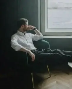 Un hombre sentado en un sillón junto a la ventana mira hacia afuera con gesto pensativo, envuelto en un silencio que refleja la sensación desconcertante de he dejado a mi pareja y le da igual, cuando las expectativas emocionales no encuentran la reacción que se esperaba.