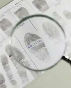 Una lupa amplía una huella dactilar en un documento de análisis, donde aparece la palabra “narcisista”, una metáfora visual que invita a observar más de cerca los rasgos y señales del narcisista vulnerable, cuya fragilidad emocional suele esconderse tras una apariencia defensiva.