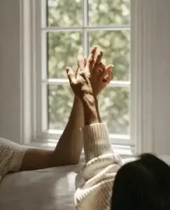 Dos manos se entrelazan suavemente frente a una ventana iluminada por la luz natural, una escena íntima que sugiere cercanía emocional y refleja las sutiles señales de atracción entre amigos.