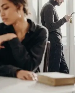 Pareja distanciada en una habitación luminosa, con una mujer triste en primer plano y un hombre mirando el móvil al fondo, mostrando cómo se comporta cuando un hombre no te quiere.